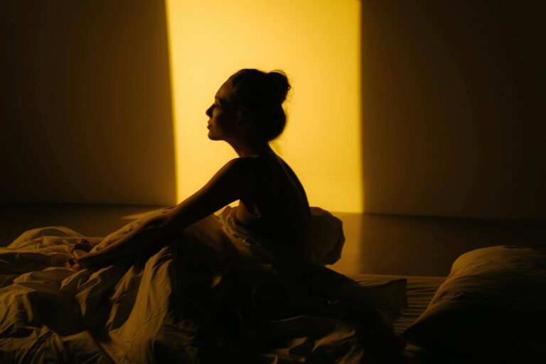 Mujer sentada en la cama, envuelta en sábanas y en penumbra, iluminada por una luz tenue. Representa el aislamiento emocional y el sufrimiento interno característicos del trauma relacional.