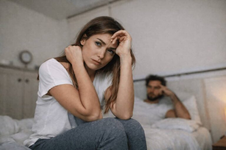 Mujer preocupada sentada al borde de la cama mientras su pareja permanece distante al fondo, reflejando la soledad emocional de quien se pregunta si su pareja es narcisista