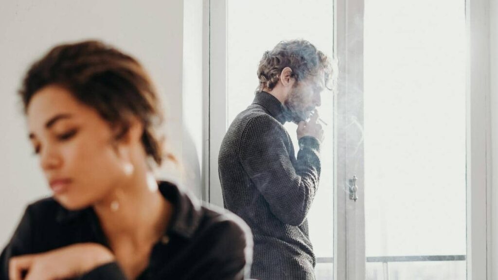 Hombre pensativo fumando mientras su pareja aparece desenfocada al fondo, representando la desconexión emocional en una relación con un narcisista